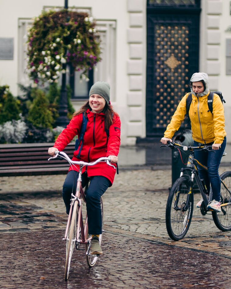 Дві дівчини в теплому одязі їдуть велосипедами по замощеній бруківкою пішохідній зоні. Перша з них широко посміхається. Погода дощова