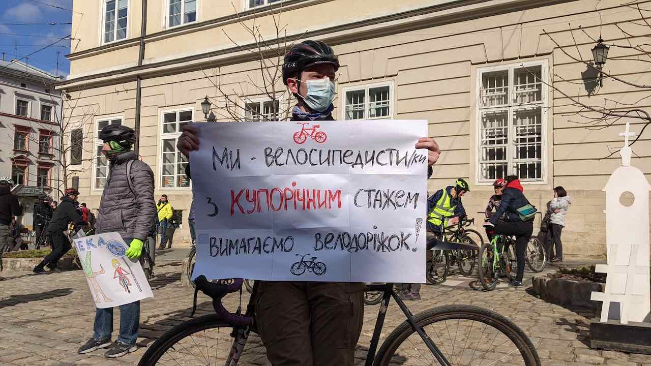 Чоловік у шоломі, спираючись на велосипед, тримає плакат з написом "Ми — велосипедисти/ки з купорічним стажем вимагаємо велодоріжок!"